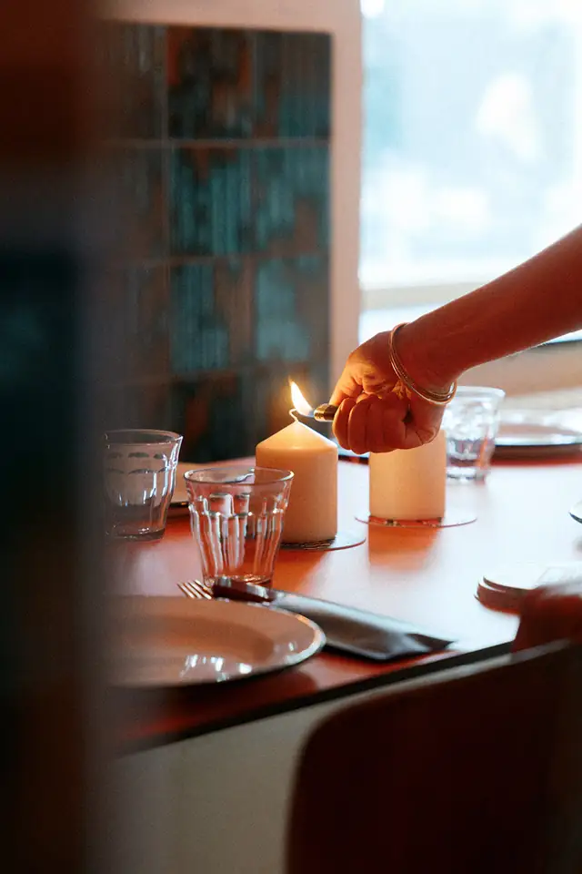 Een hand steekt een kaars aan op een gedekte tafel bij het raam