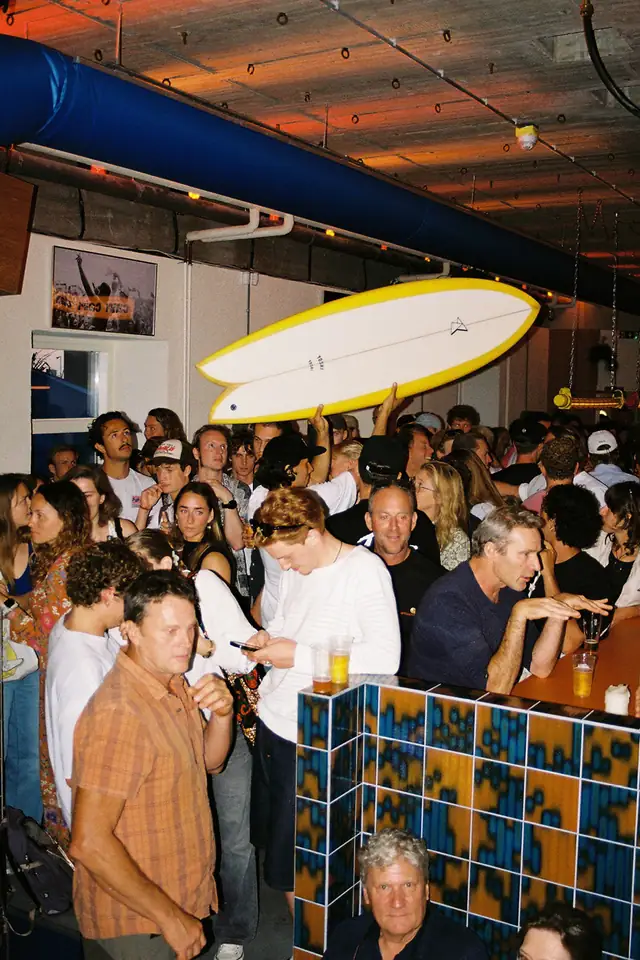 Een volle zaal gasten bij de bar, met een gele surfplank boven het publiek