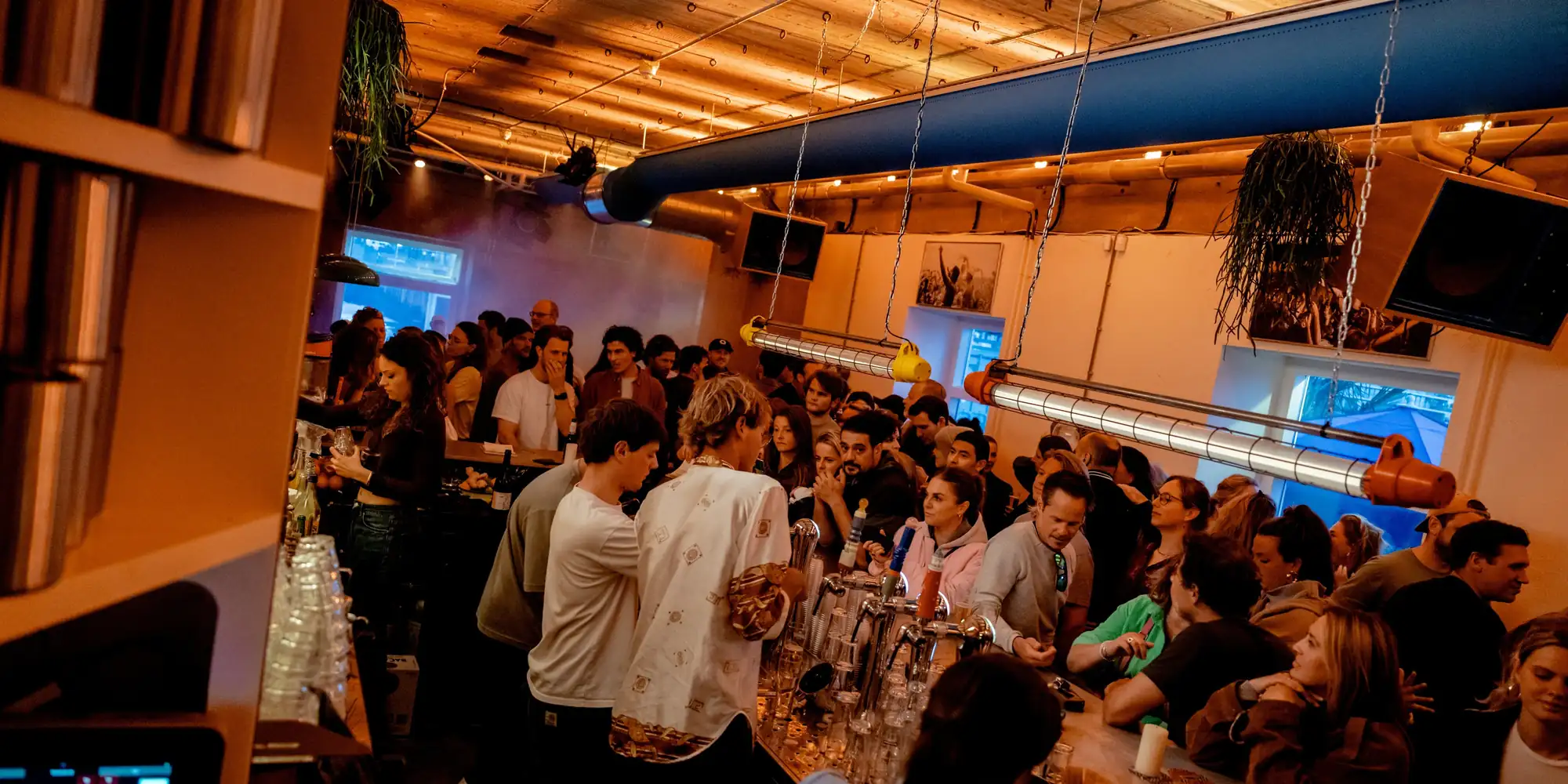 Een volle zaal gasten aan lange tafels onder het oranje licht van Caipi Café