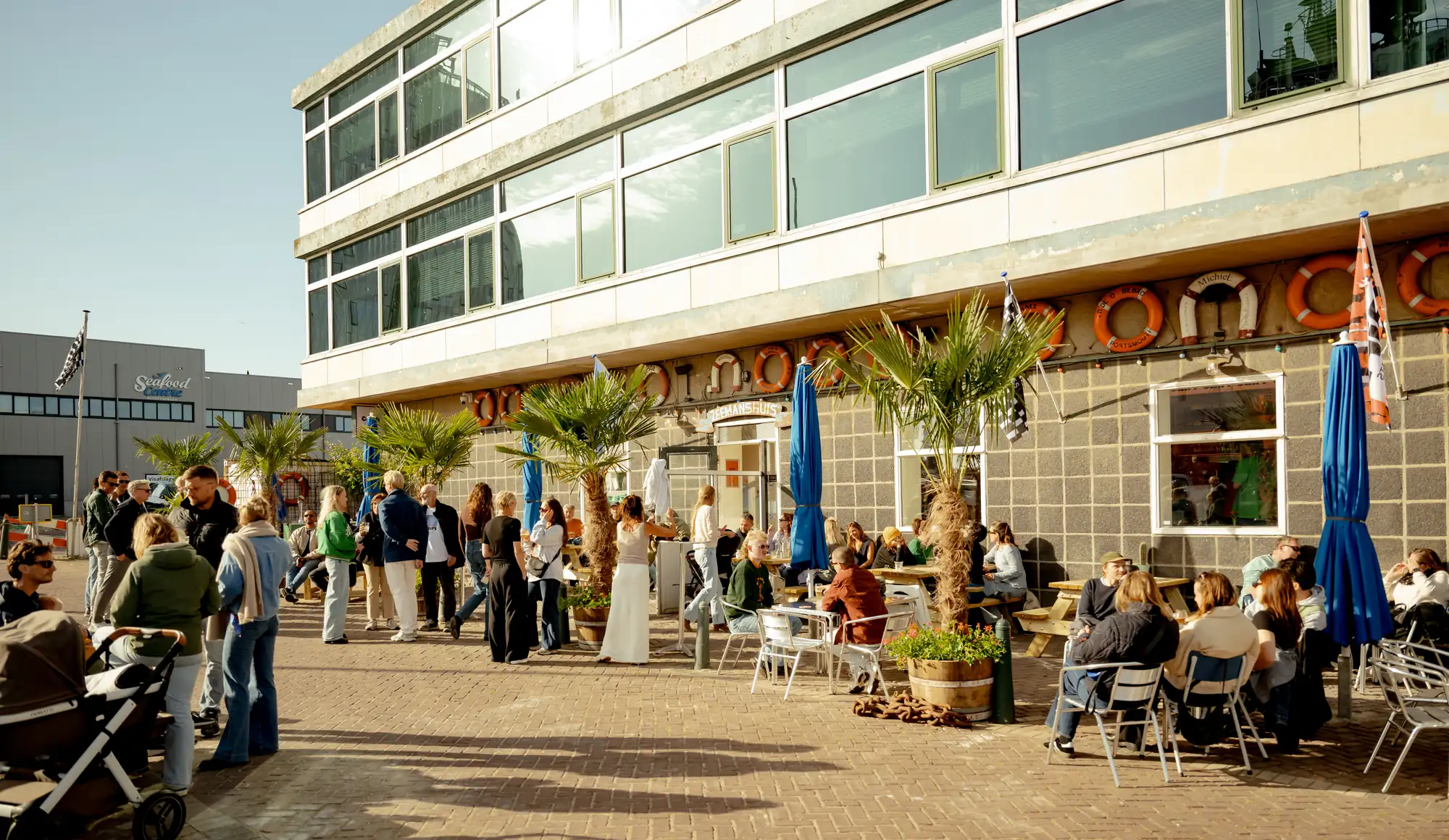 Het terras van Caipi Café in de middagzon, met palmen, blauwe parasols en de gevel van het Zeemanshuis met rode reddingsbanden