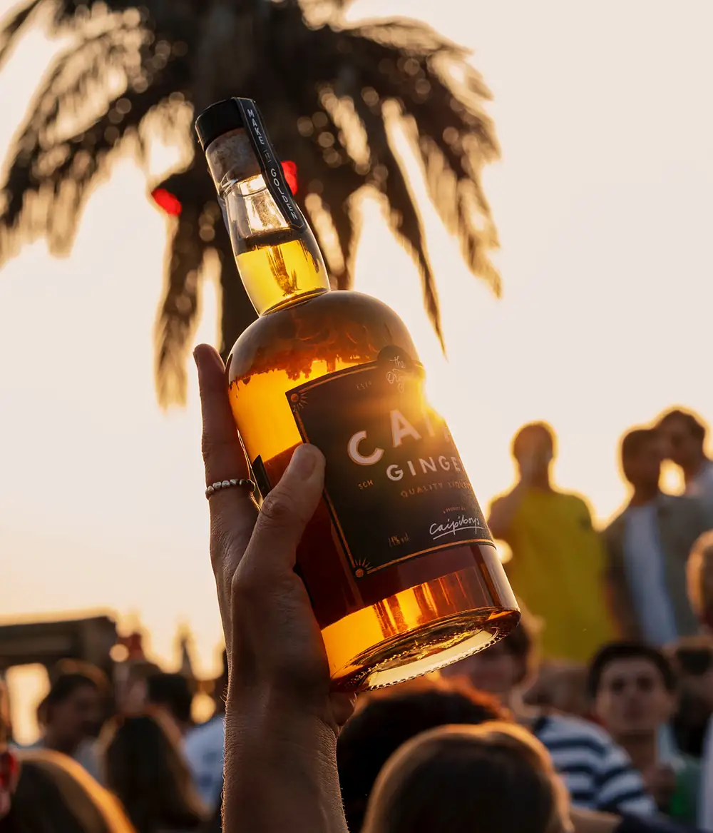 A hand holds up a bottle of Caipi Ginger against a palm tree and the evening sun, with a crowd below