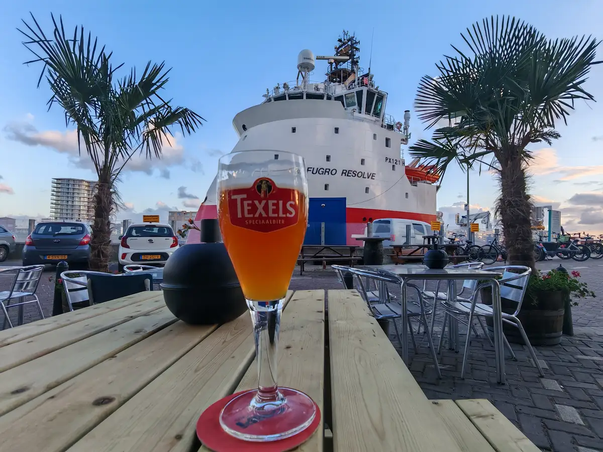 Een glas Texels bier op een picknicktafel op het terras, met palmen en een schip in de haven erachter